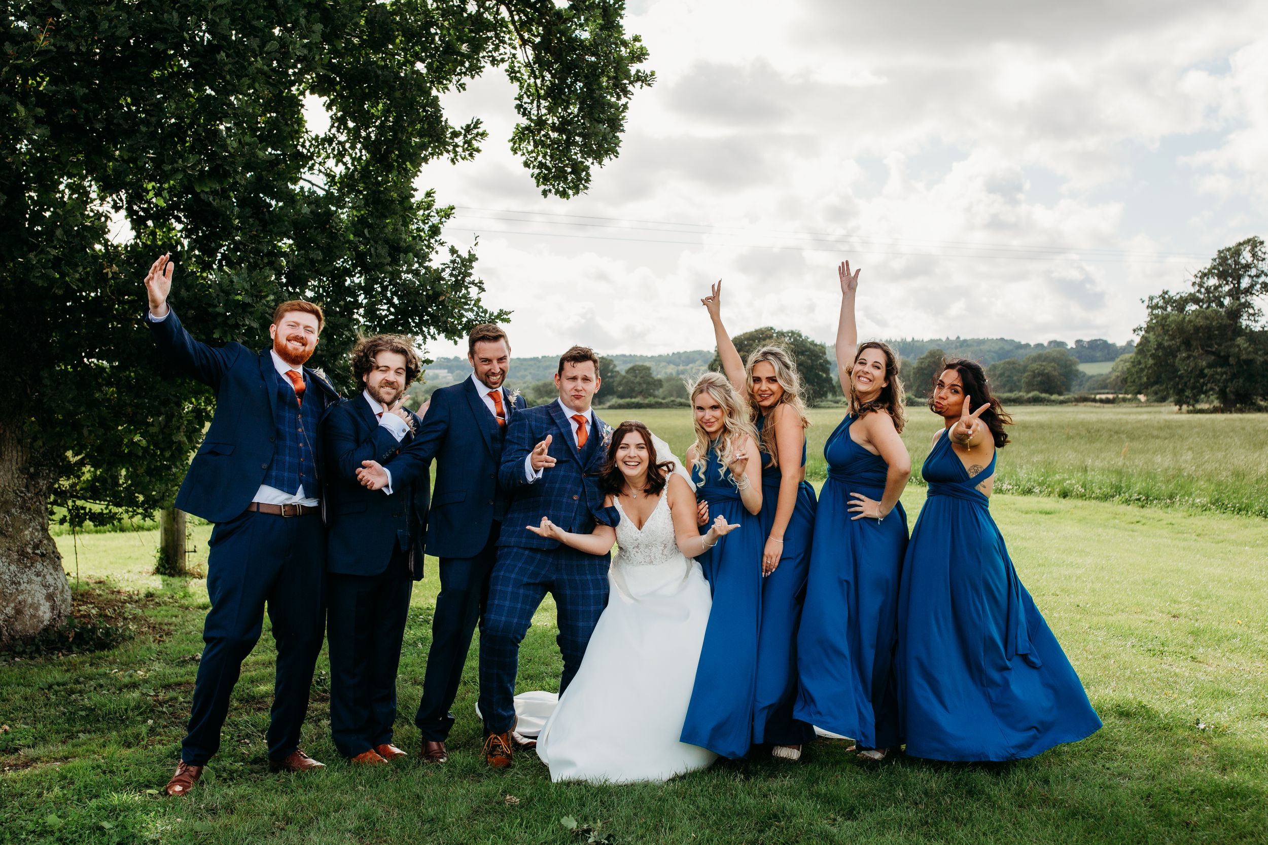 Wedding party celebrates on a grassy hillside with bridesmaids in blue dresses.