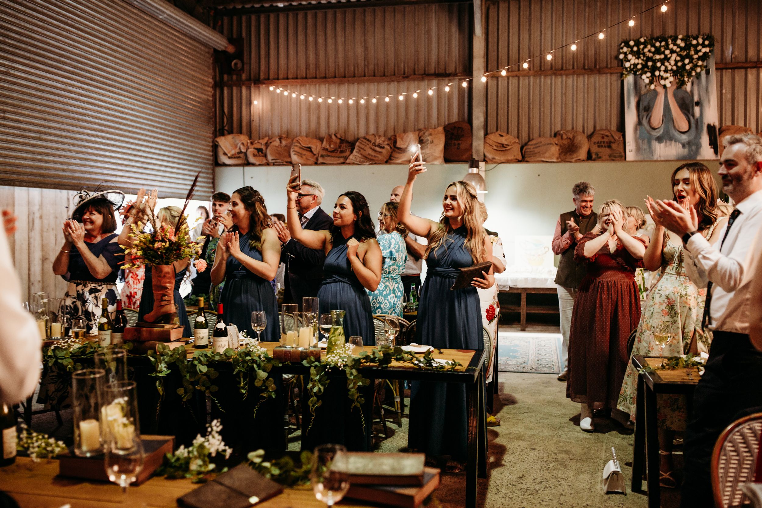 Wedding reception guests toast and celebrate at tables decorated with flowers and string lights.