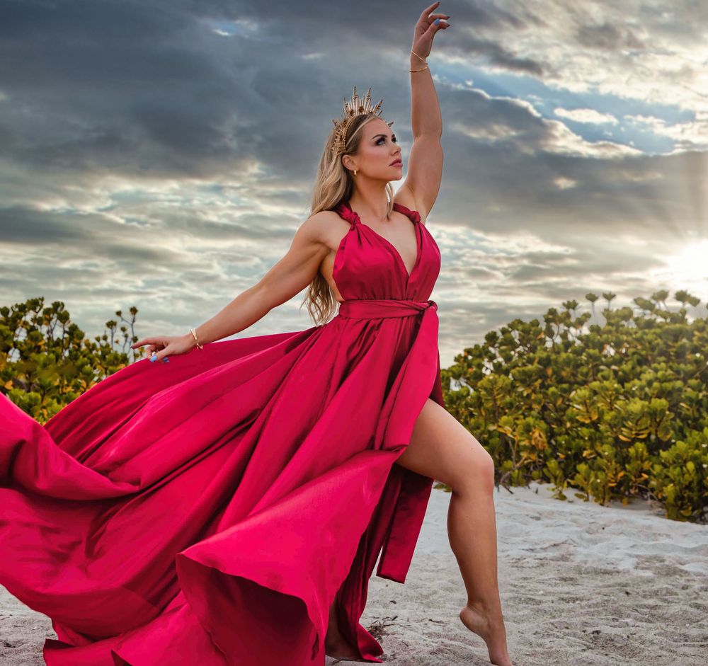Red dress flows dramatically during beach sunset photoshoot with cloudy sky.