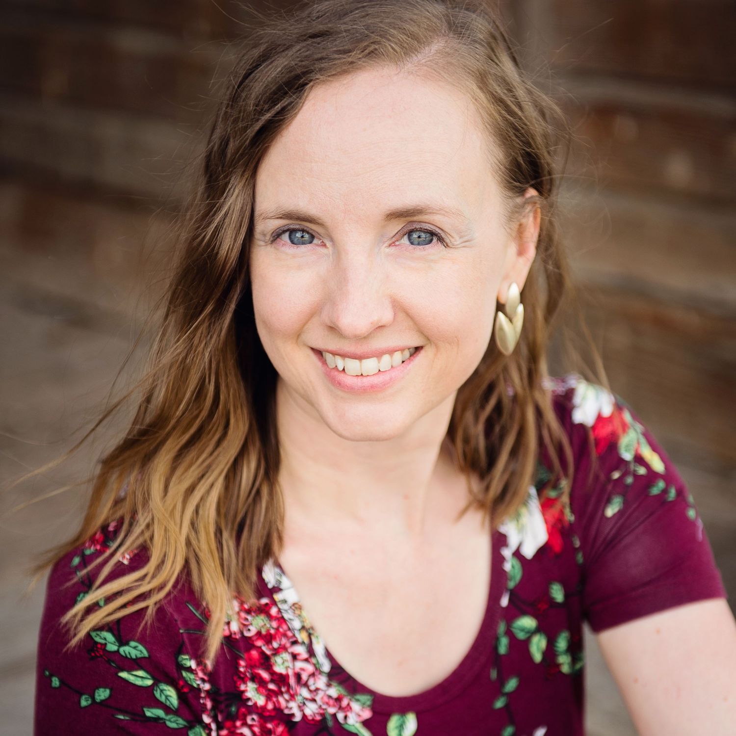 A professional headshot features a burgundy floral top against a rustic wood background.