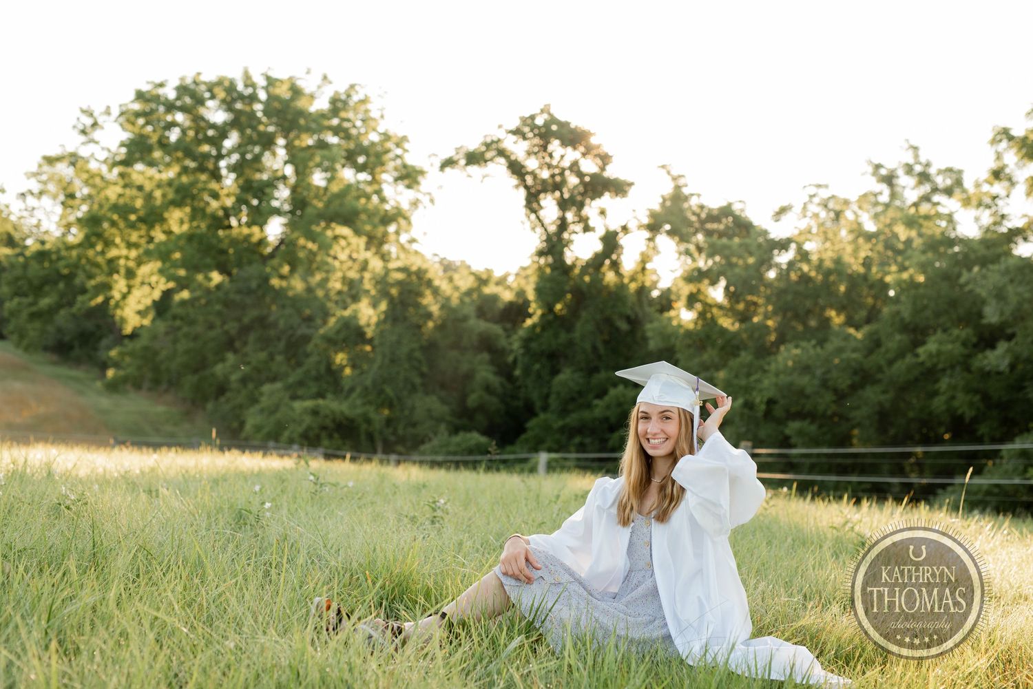 Graduate sits casually in sunny field wearing white graduation attire with trees in background.