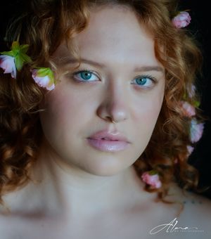 Portrait with striking blue eyes and curly auburn hair adorned with delicate pink flowers against dark background.
