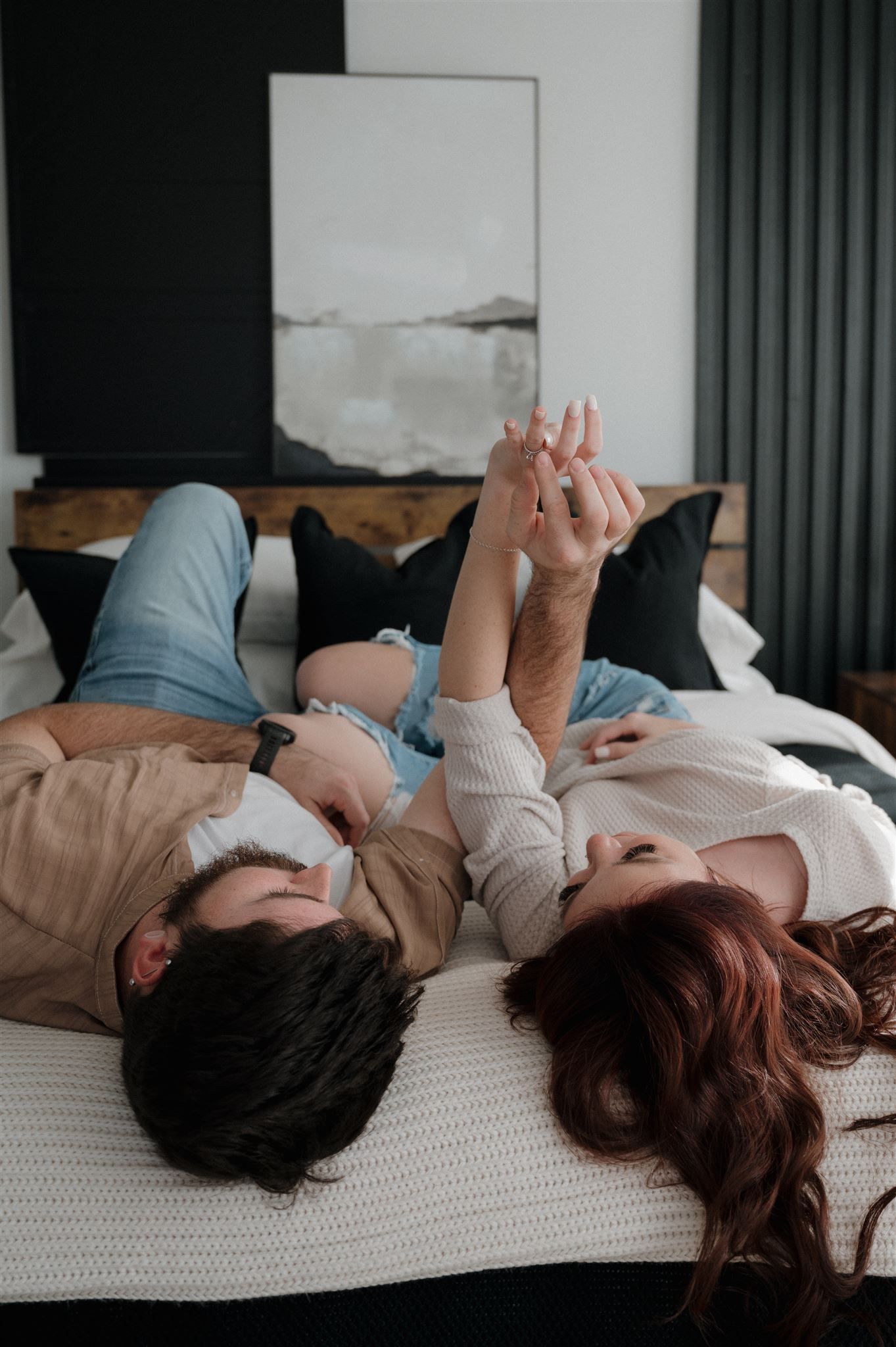 Couple holding hands while lying on bed with minimalist decor in background.