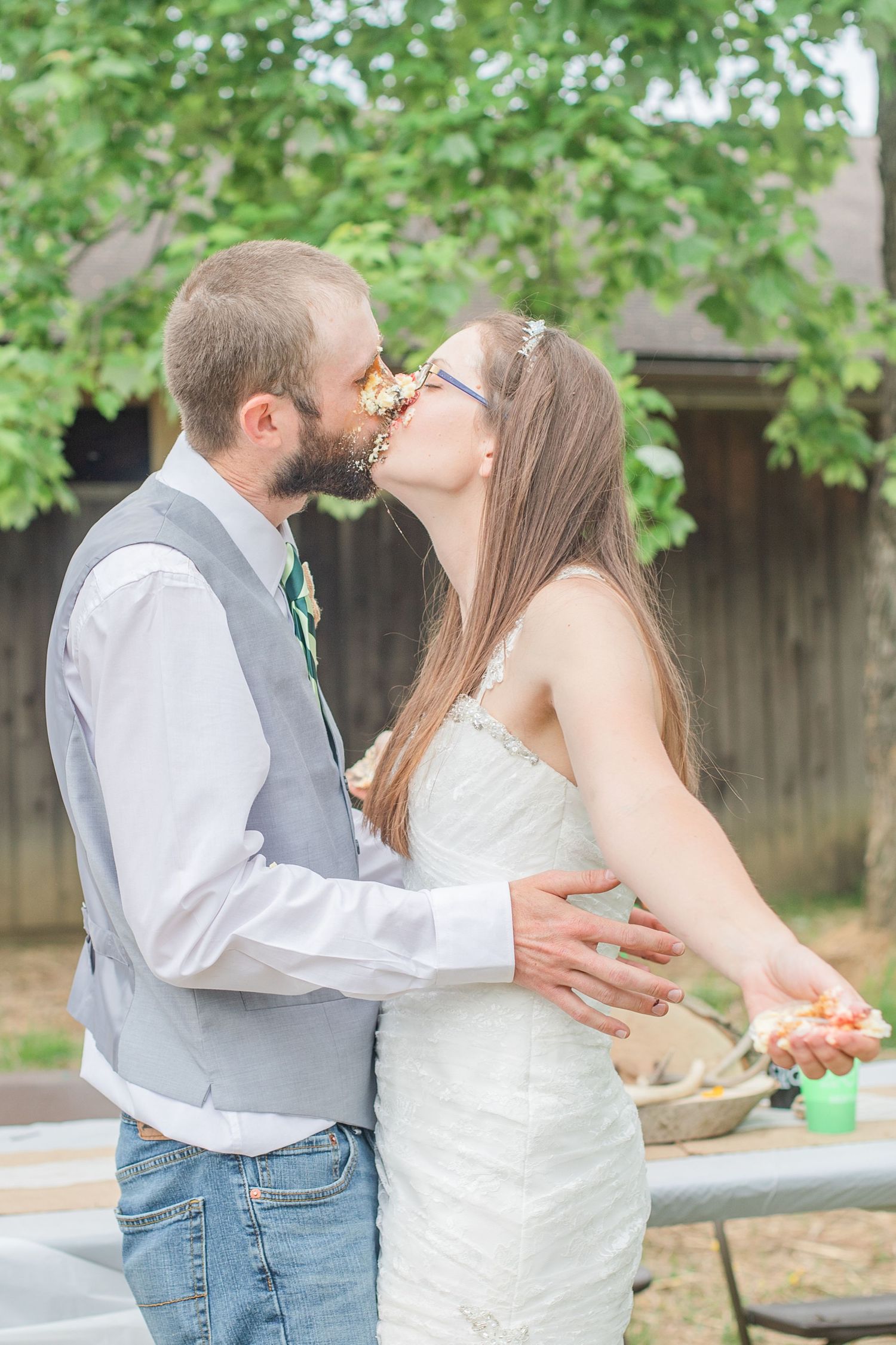 A couple shares a romantic kiss outdoors with greenery in the background during their wedding celebration.