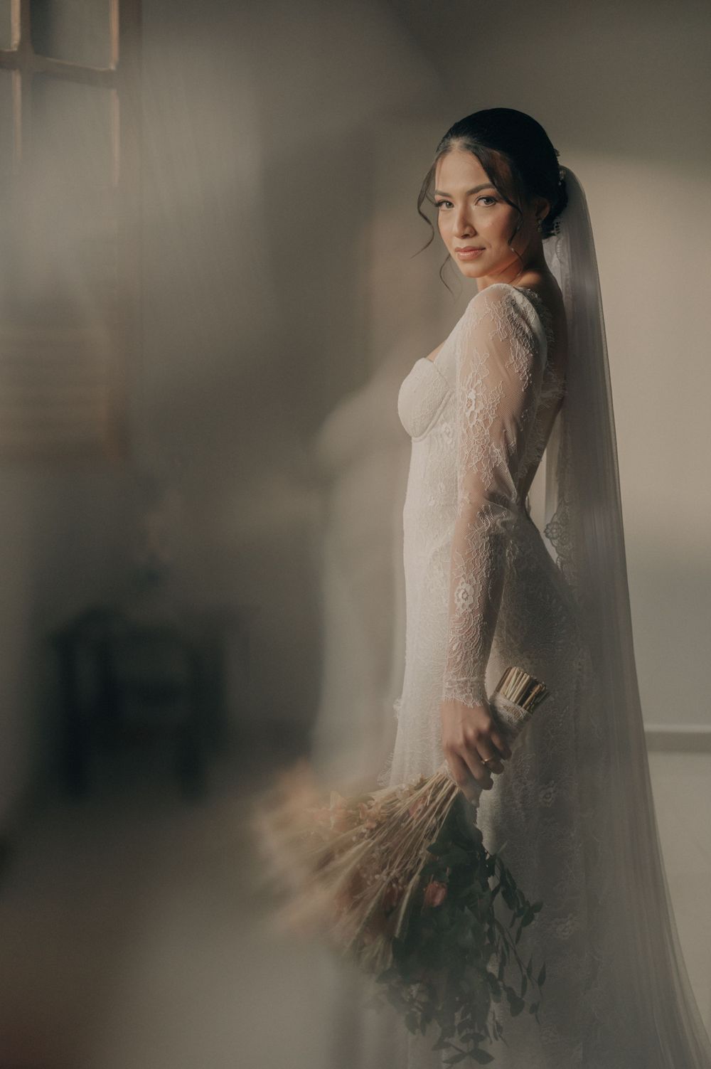 A bride in a long-sleeved lace gown holds a dried flower bouquet in moody lighting.