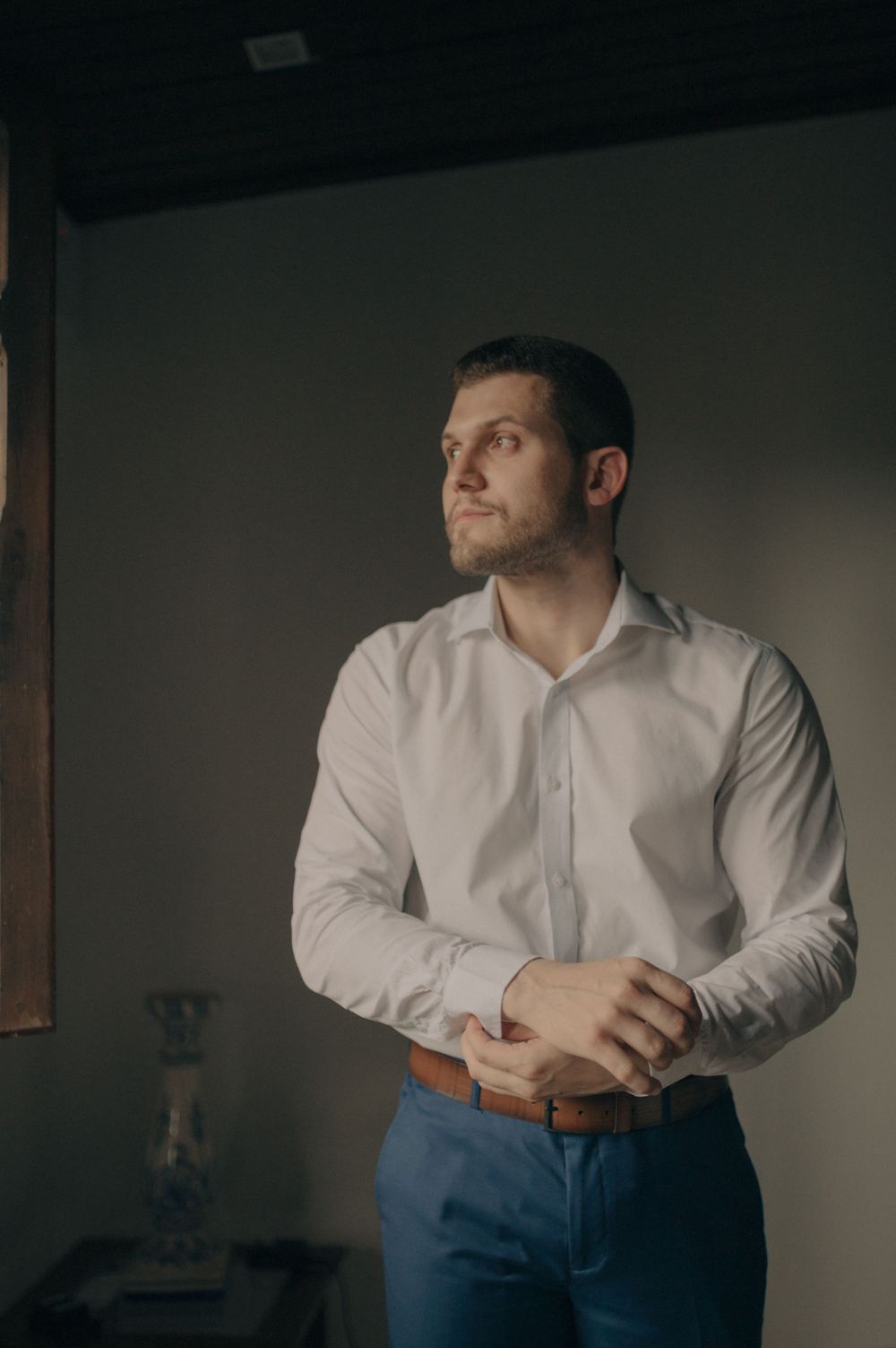 A person in a crisp white dress shirt and blue slacks stands in contemplative pose by a window.