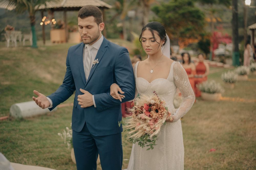 A couple walks together outdoors during their wedding ceremony with coral-dressed attendants visible behind.