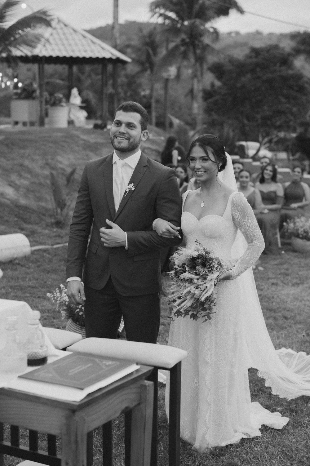 A black and white photo of a wedding ceremony outdoors with palm trees and a rustic podium setup.