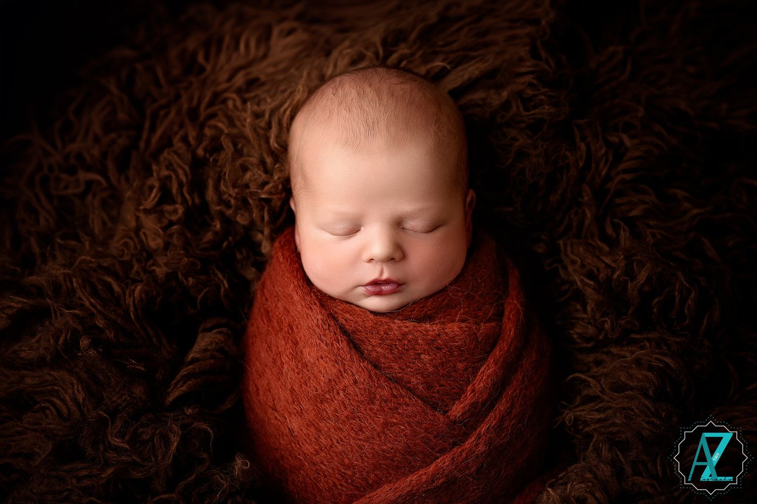 Newborn photography featuring sleeping infant wrapped in rustic red blanket against dark textured backdrop.