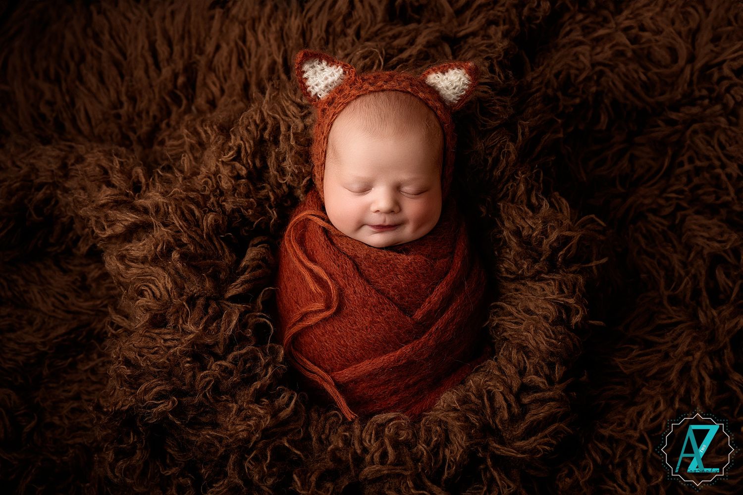 Sleeping newborn wrapped in rust-colored wrap wearing fox ears on brown fluffy texture.