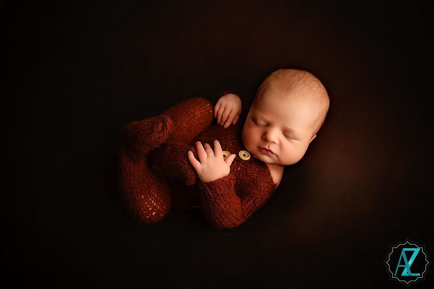 Peaceful sleeping infant in brown knit outfit with teddy bear on dark background.