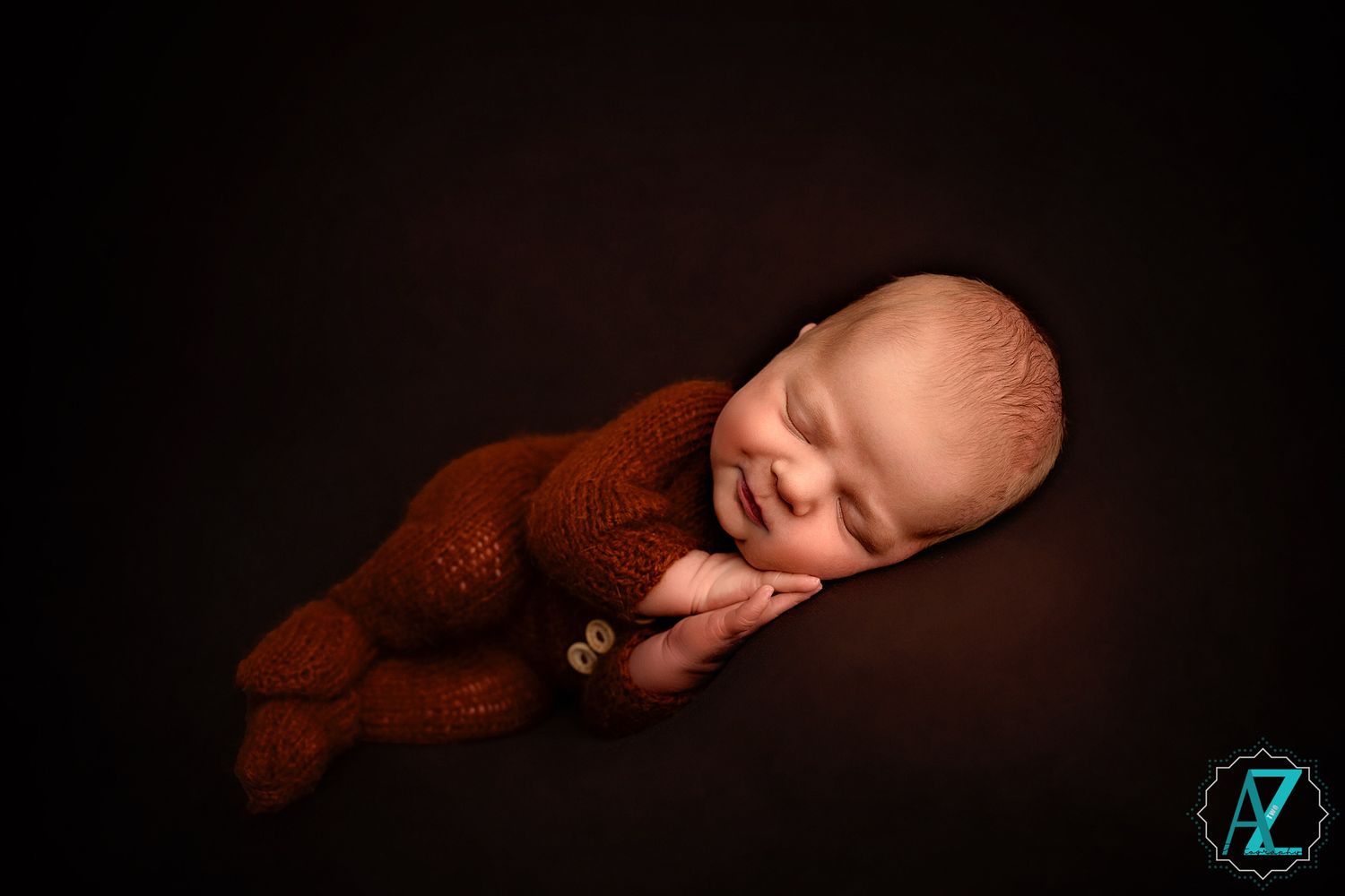 Peaceful sleeping newborn in brown knit outfit on dark moody backdrop.