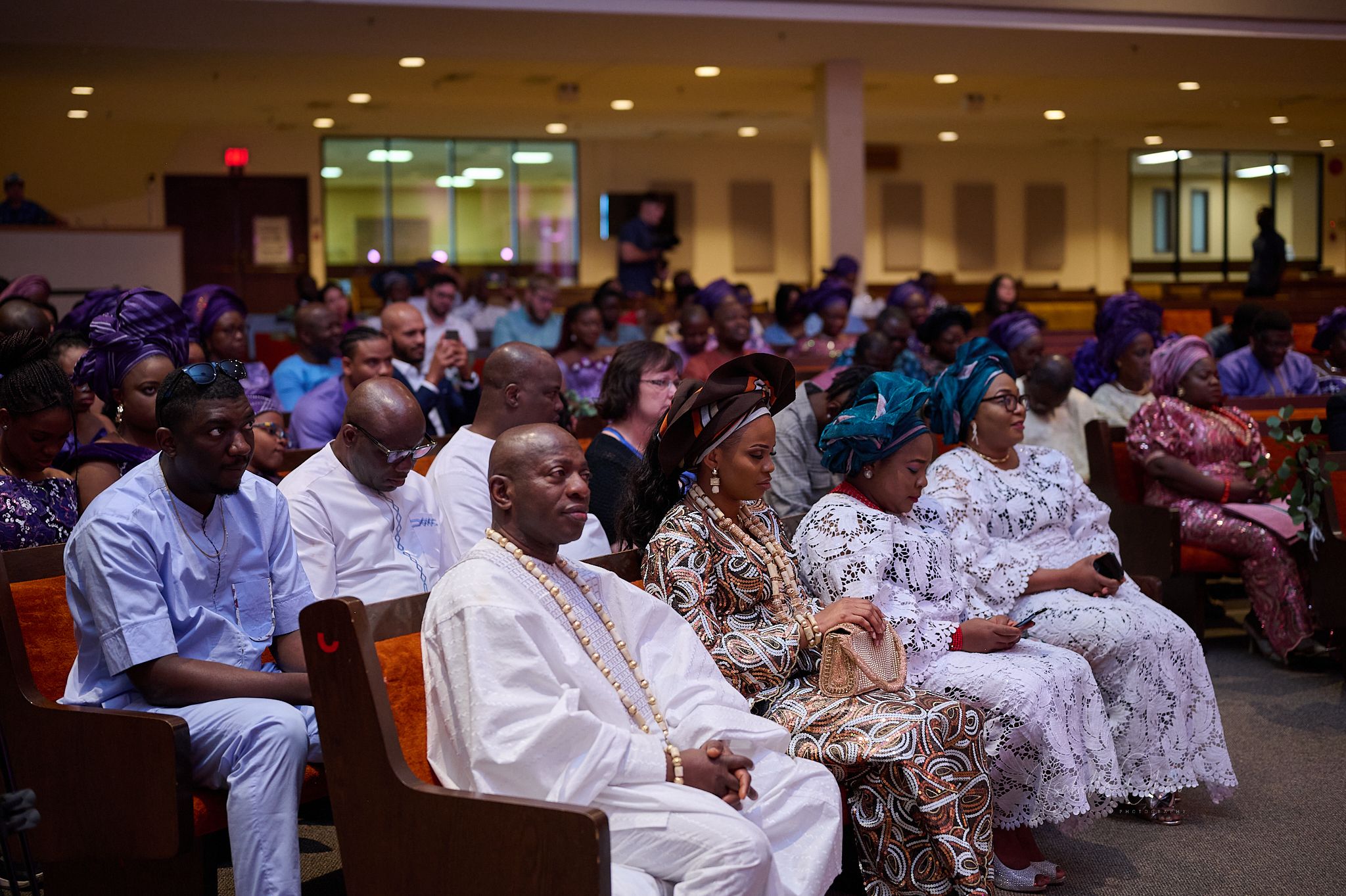 Wedding guests seated in rows during a ceremony wearing colorful formal attire in a large hall.