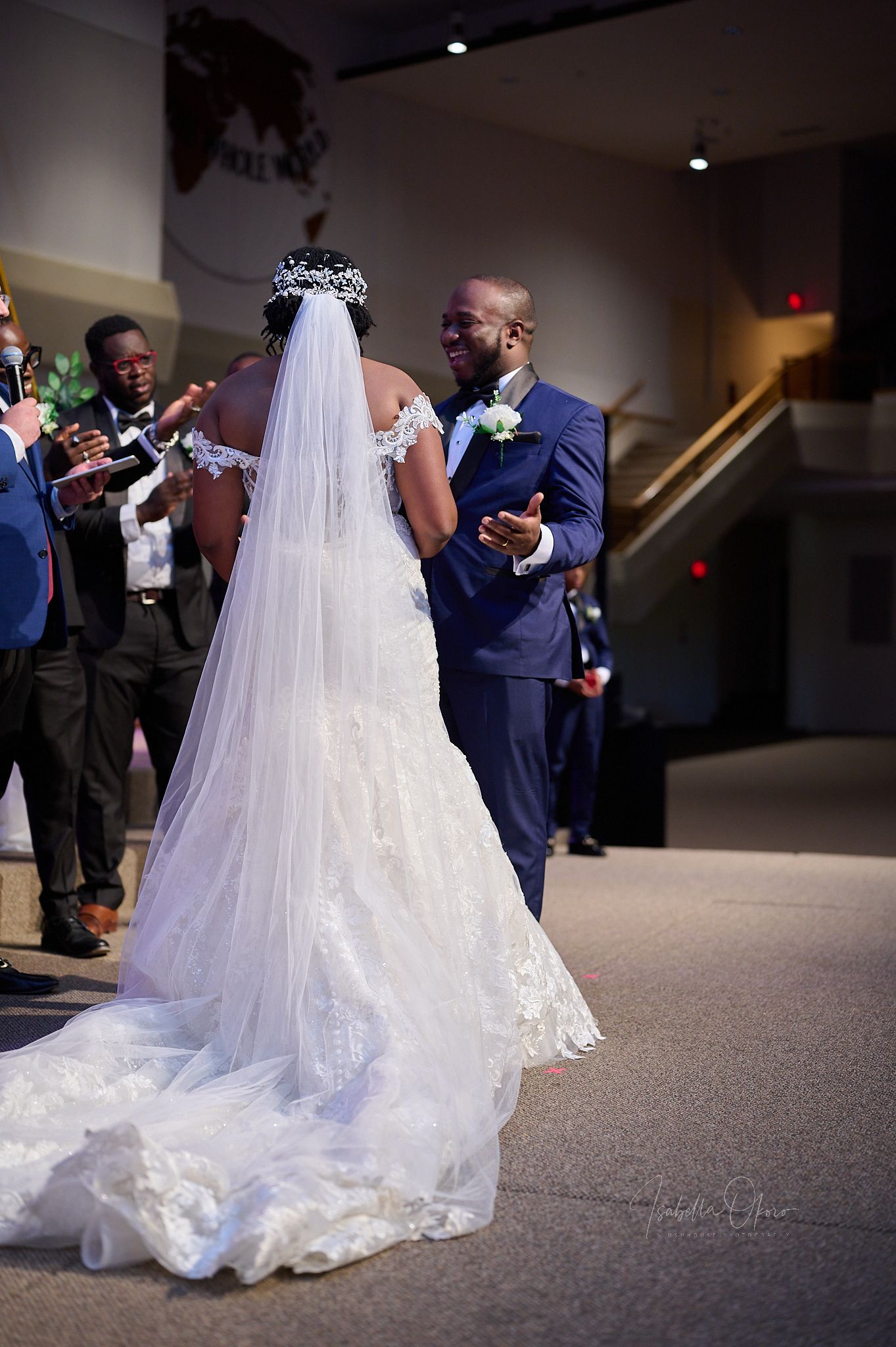 A bride in an elegant lace wedding dress with cathedral veil stands before wedding guests.