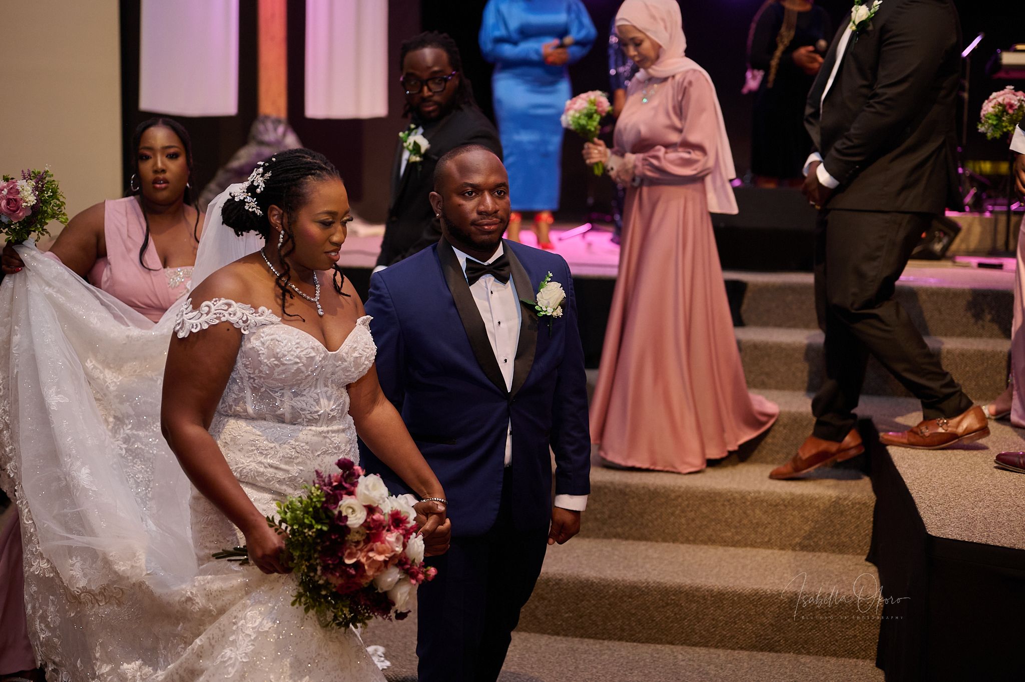 Wedding party members walk down church steps after ceremony with bride carrying pink bouquet.