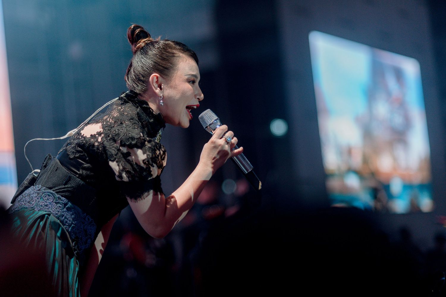 A performer sings passionately into a microphone on stage with blue lighting and video screens behind.