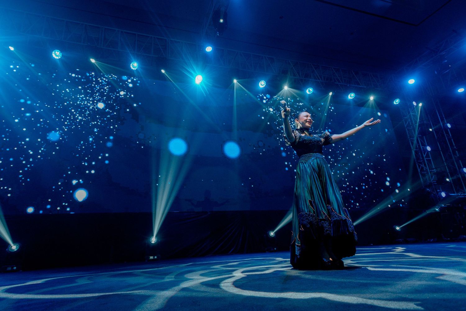 A performer in a flowing dark dress performs under ethereal blue stage lighting with sparkle effects.