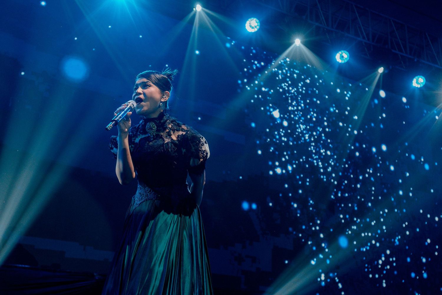 A performer sings on stage surrounded by dramatic blue lighting and sparkling visual effects.