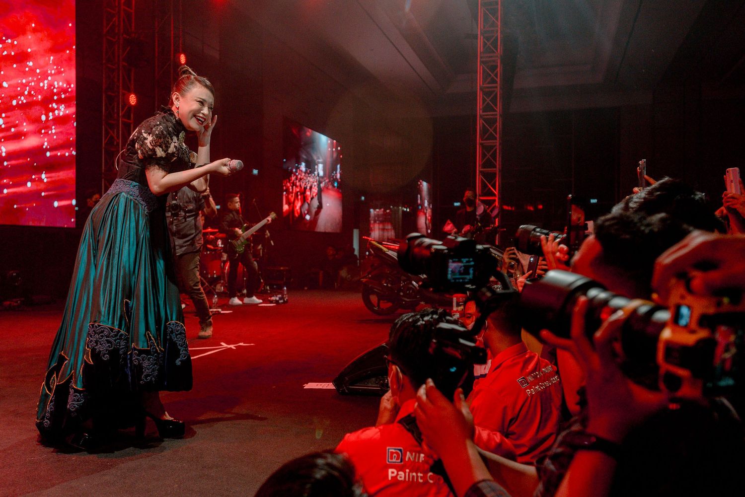 A performer in a long dress commands the stage bathed in dramatic red lighting while photographers capture the moment.