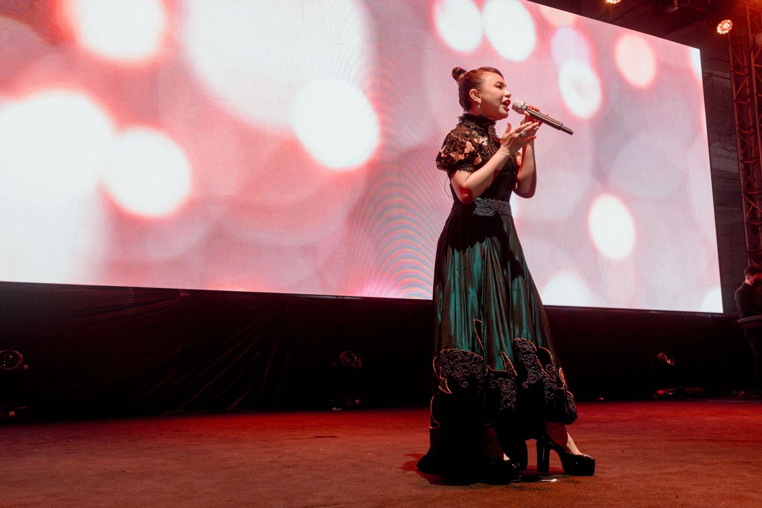 Stage performer in an emerald green gown singing against a red-lit backdrop with bokeh effects.