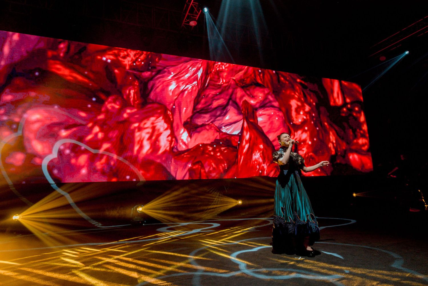 A performer stands on stage against a dramatic red backdrop with golden light beams creating an ethereal atmosphere.