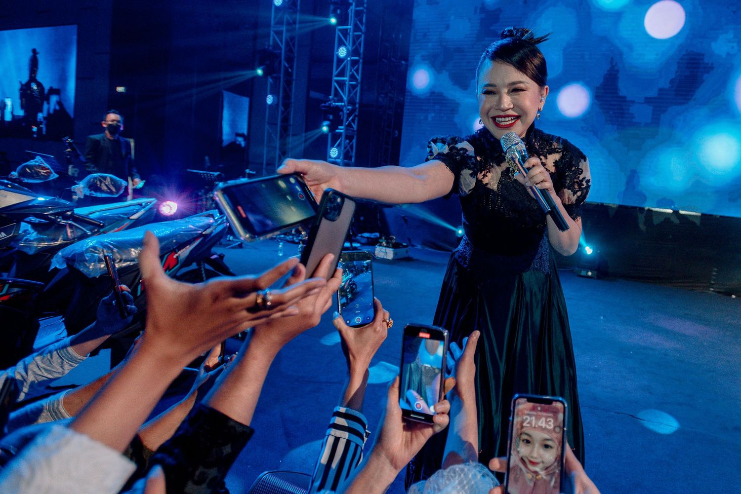 Performer in dark dress reaches out to interact with audience members holding phones at concert.