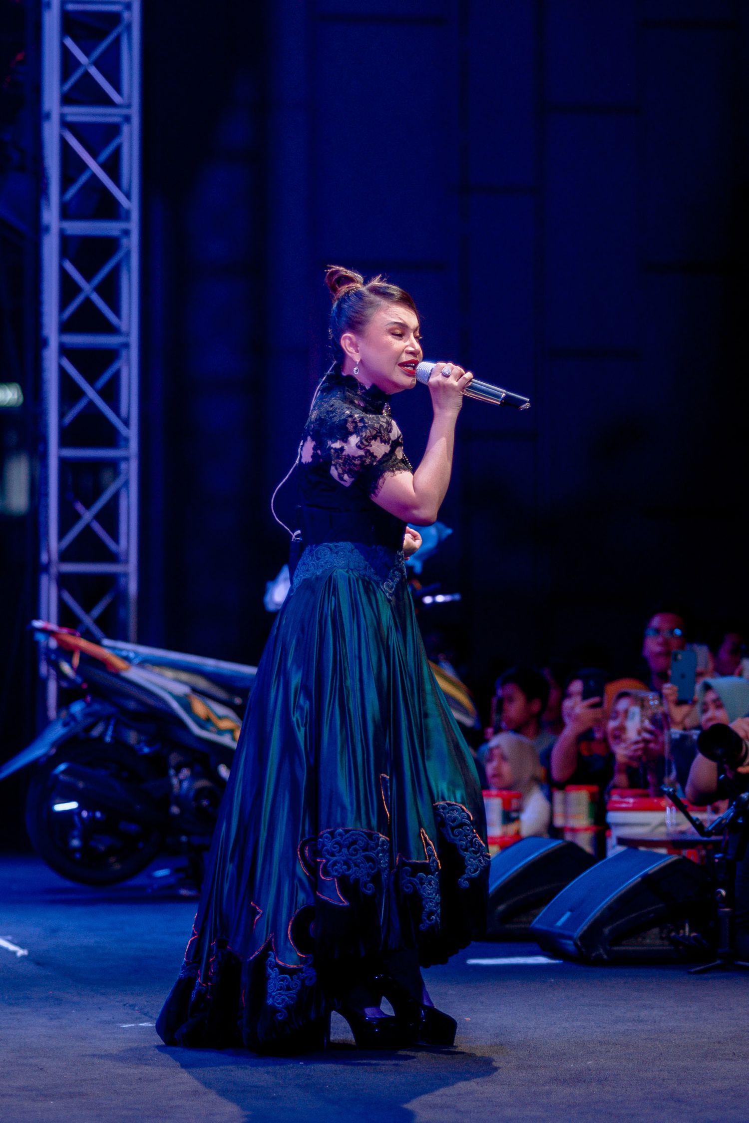 A performer in a dark blue flowing dress sings on stage under dramatic blue lighting at a concert.