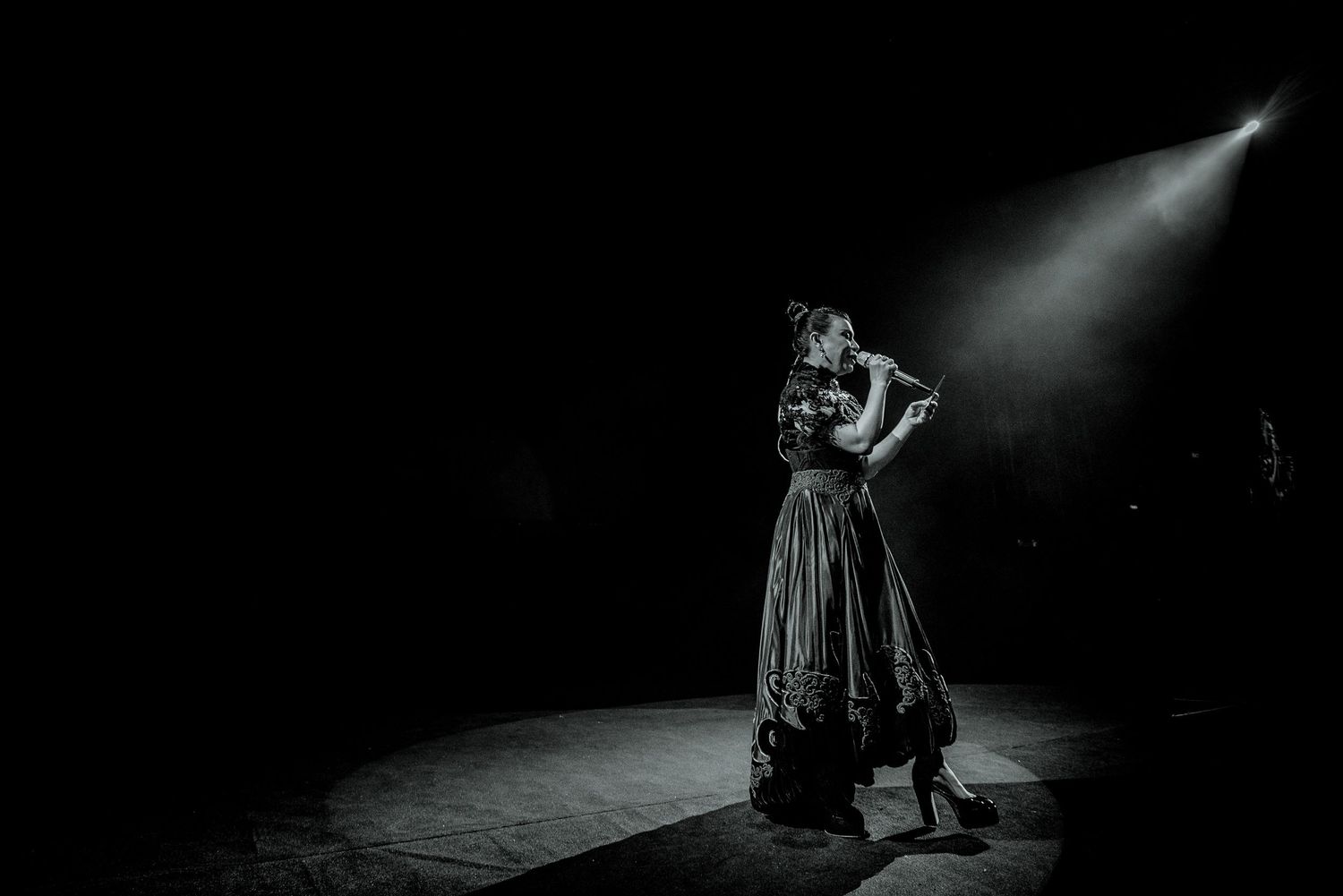 A dramatic black and white shot of a performer in a flowing dress under a spotlight on stage.