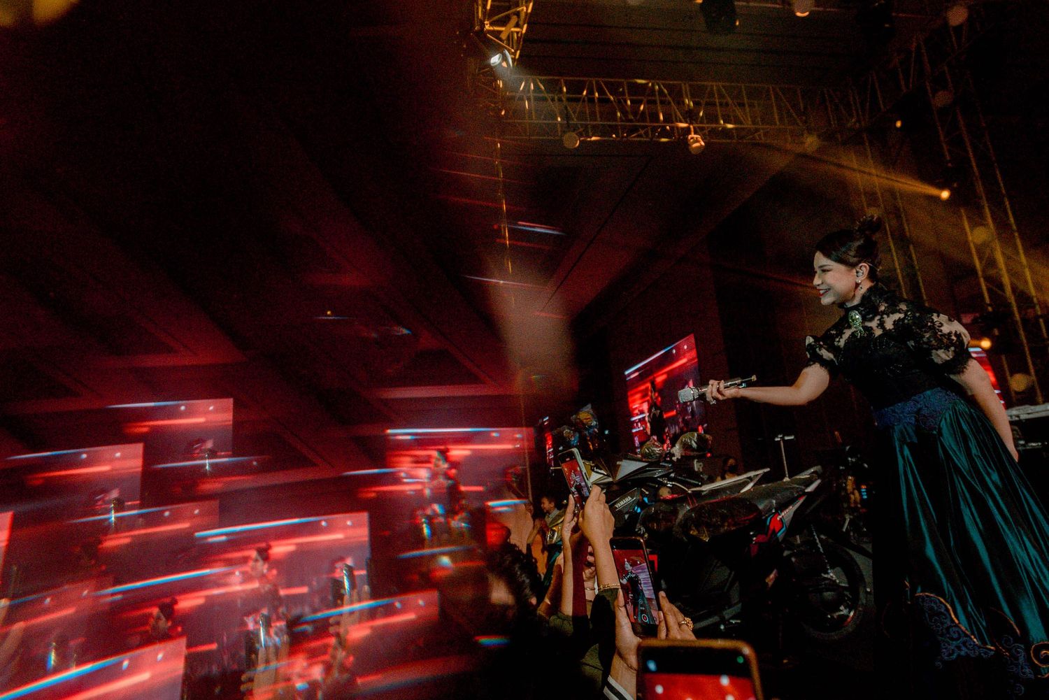A performer in a dark dress energetically performs on stage with red lighting effects in the background.