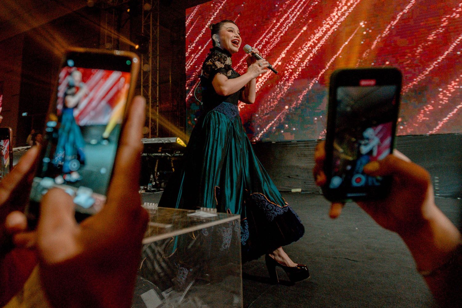 Audience members capture a performer in a teal dress through their phone screens during a live show.