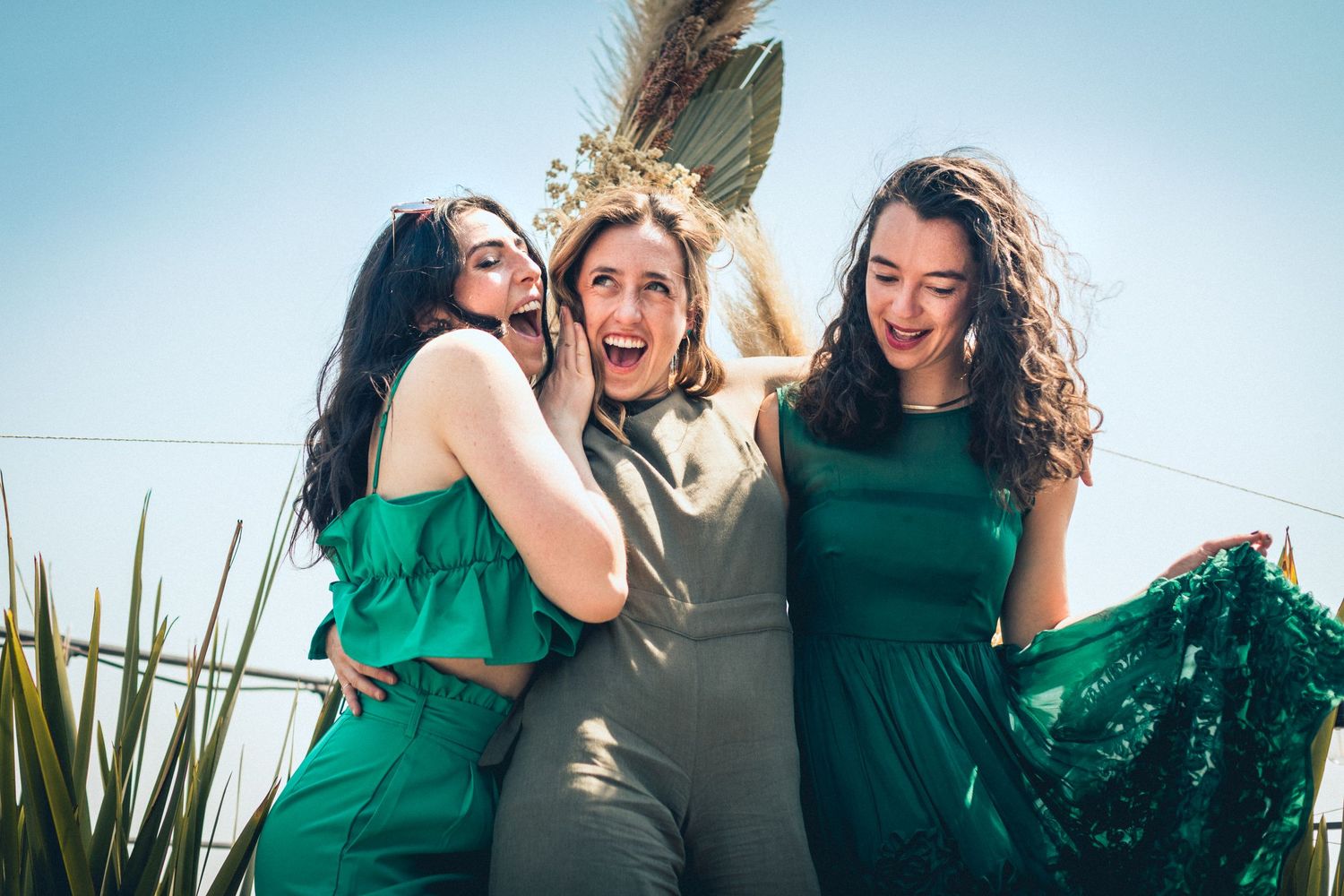 Three women in green dresses celebrating and laughing together on a sunny rooftop.