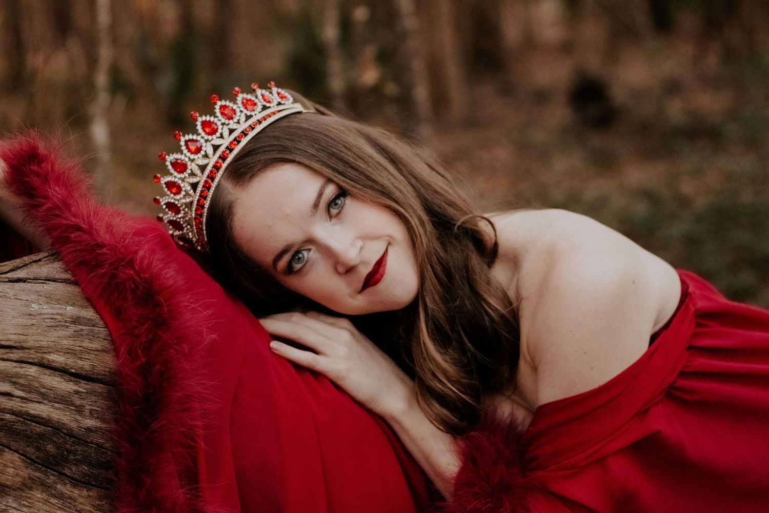 A woman in a red dress and a crown lays on a fallen tree in the woods