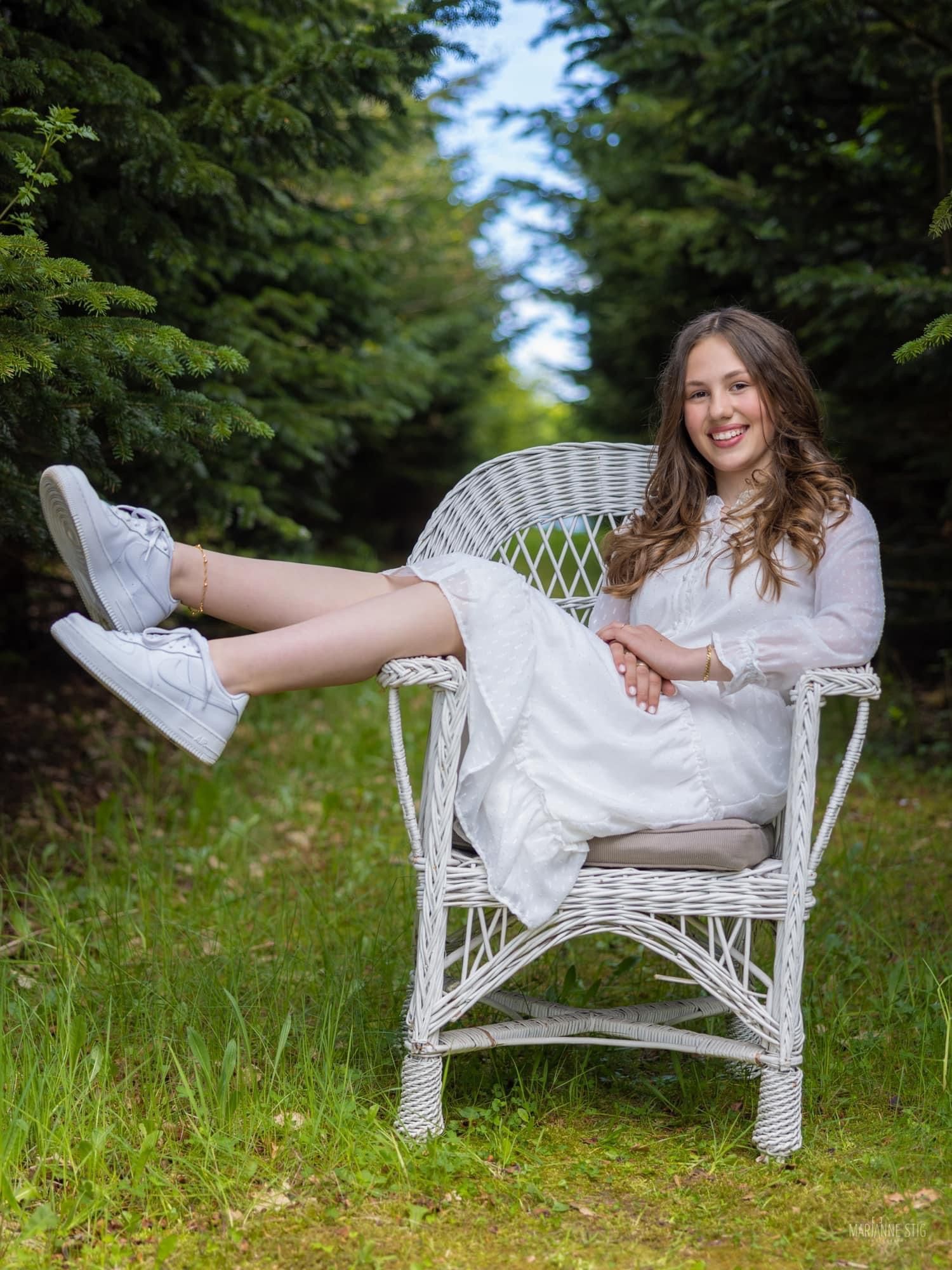 Person in white dress posing playfully on wicker chair in garden setting.