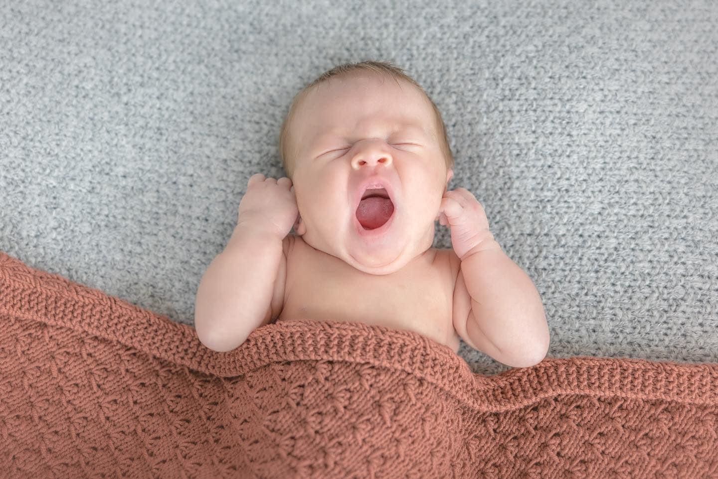 Adorable yawning baby lying on gray and coral knit blankets.