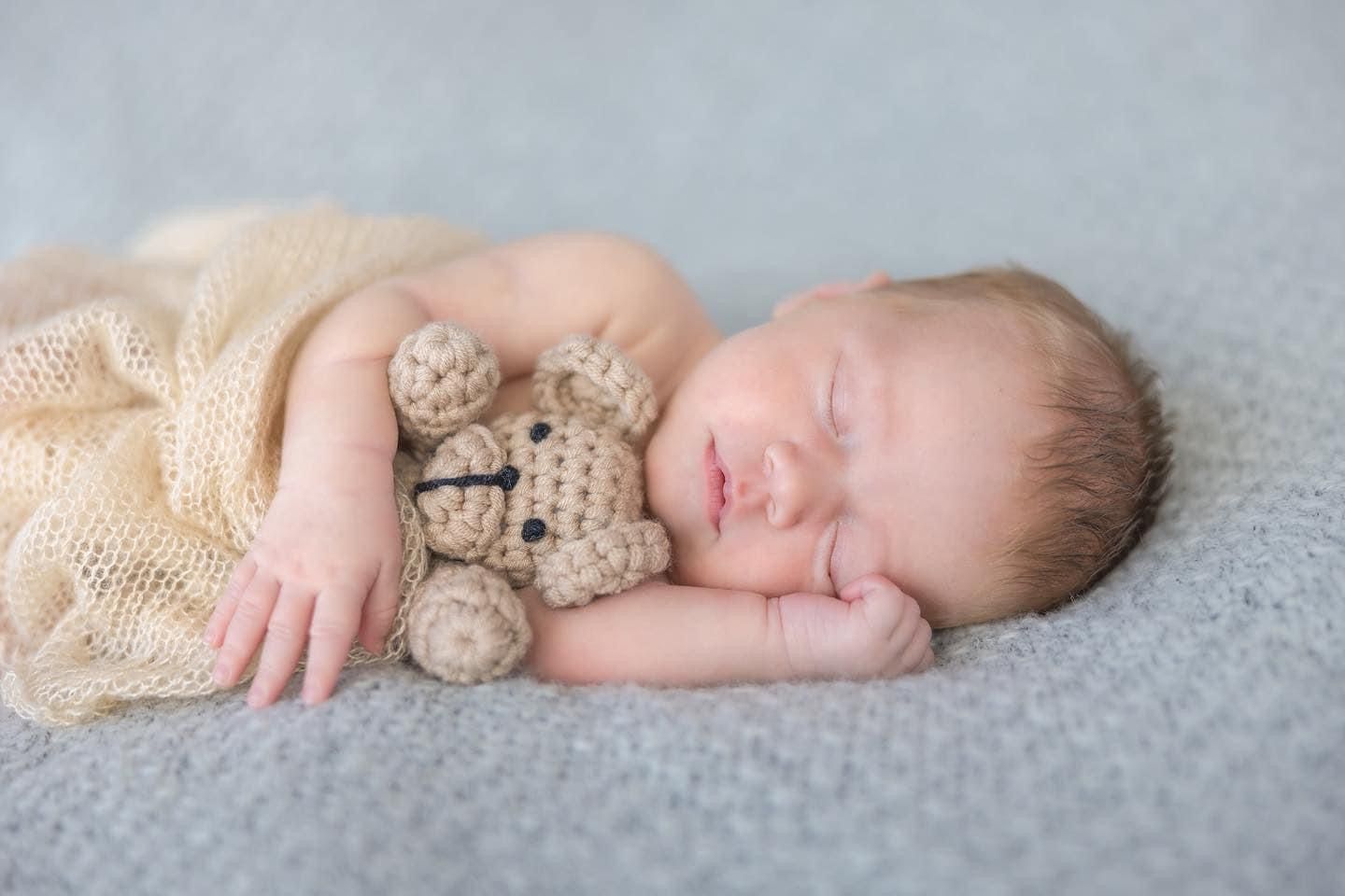 Peaceful sleeping newborn wrapped in lace holding a tiny teddy bear.