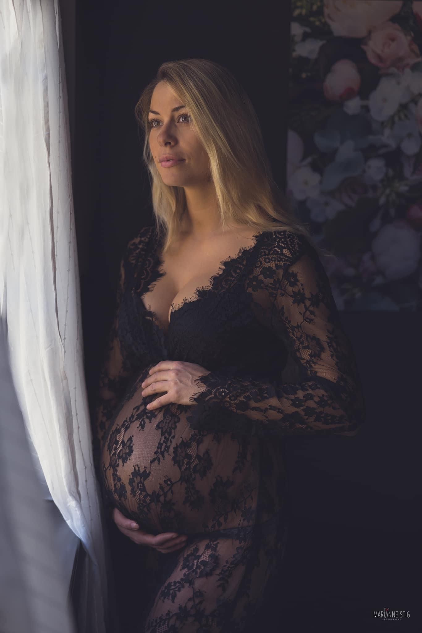 Artistic maternity portrait in black lace dress by window with dramatic lighting.