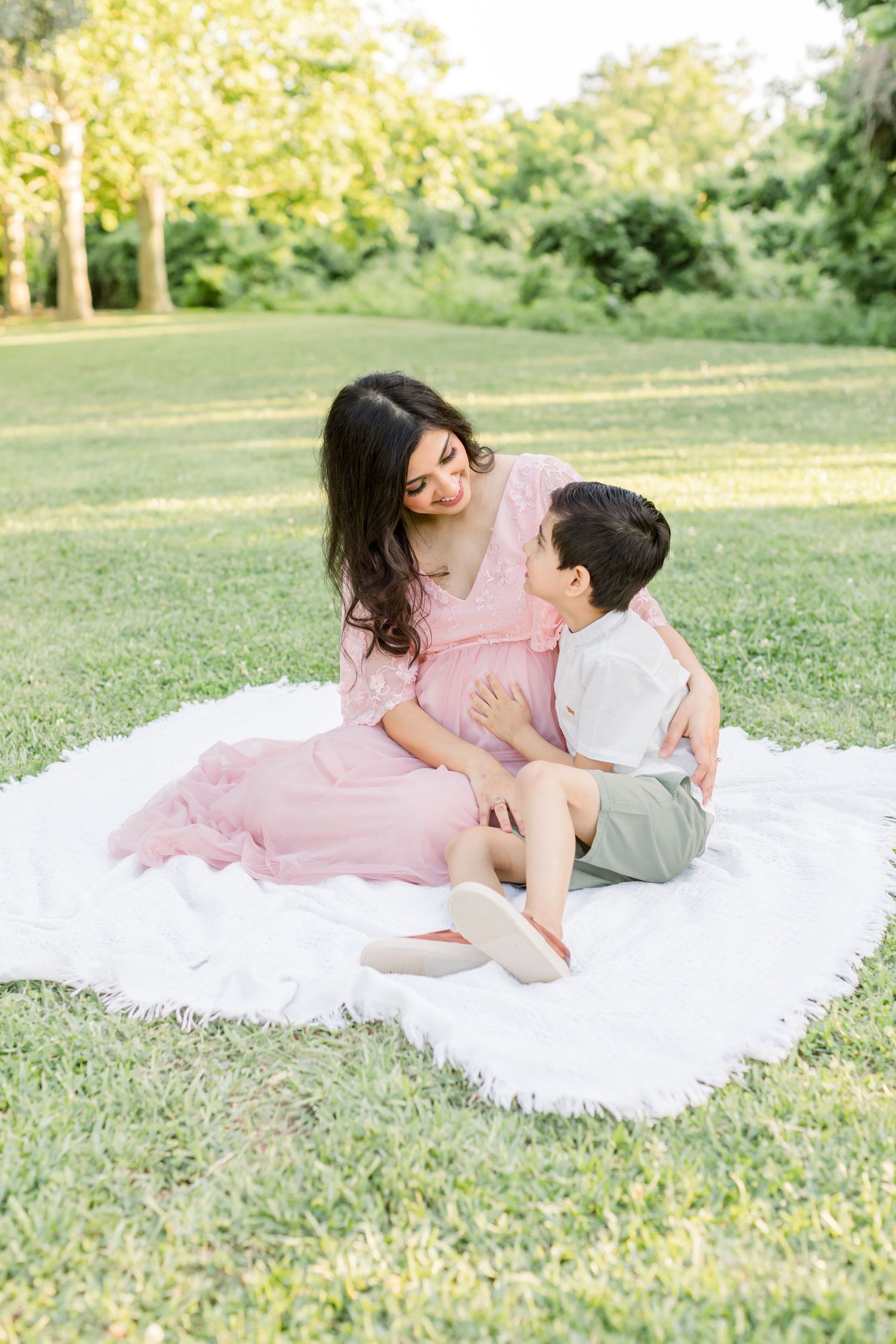 An intimate maternity moment captured on a white blanket in a sunny outdoor setting.