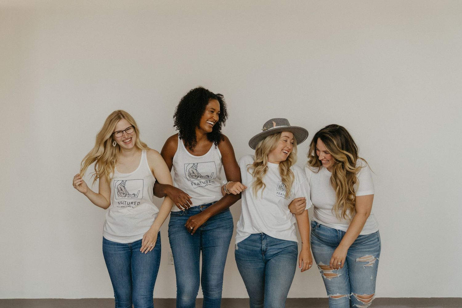 Four friends in matching white tops and jeans share laughs against white wall.