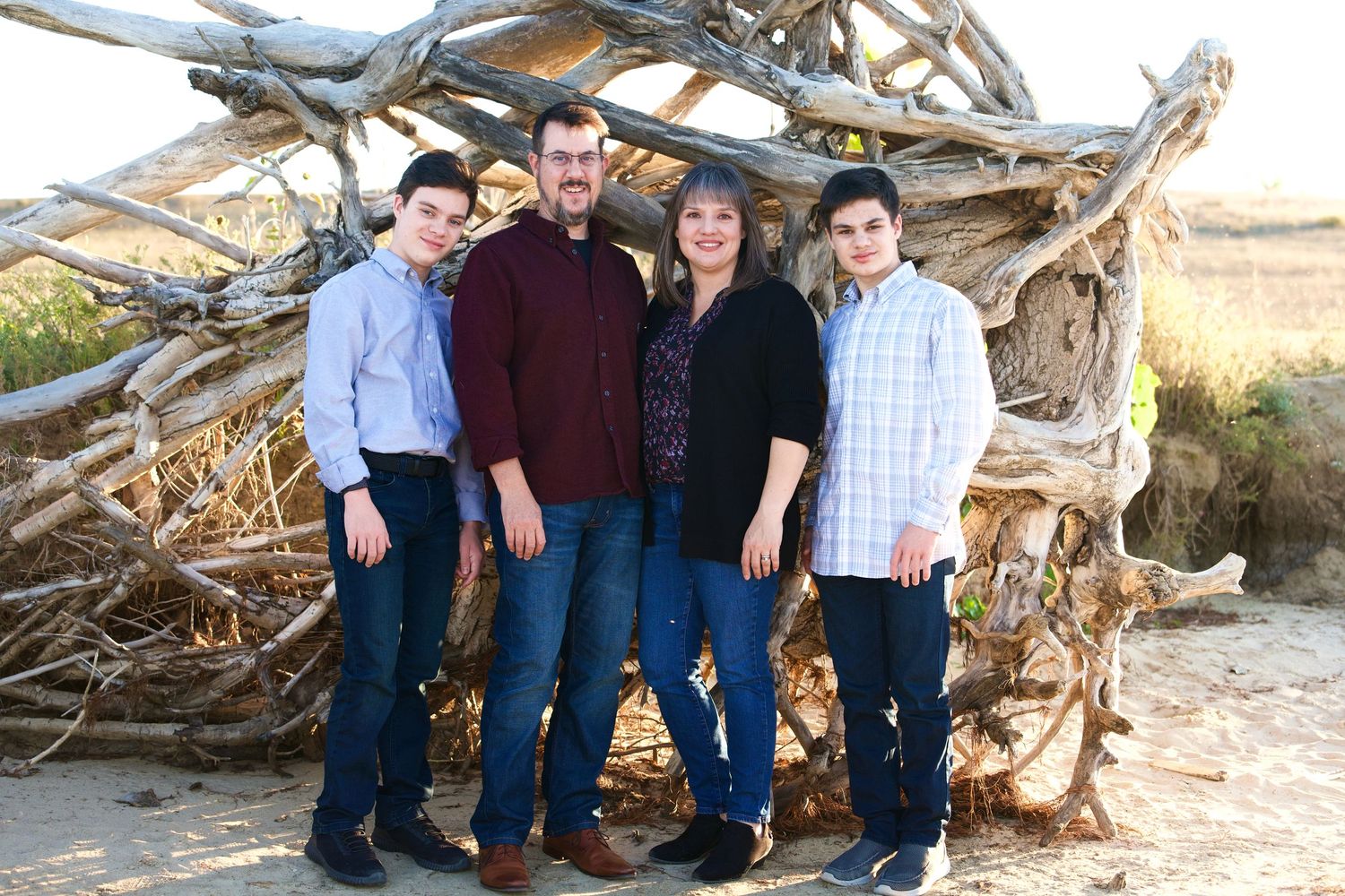 Family group portrait against rustic driftwood structure in casual coordinated outfits.