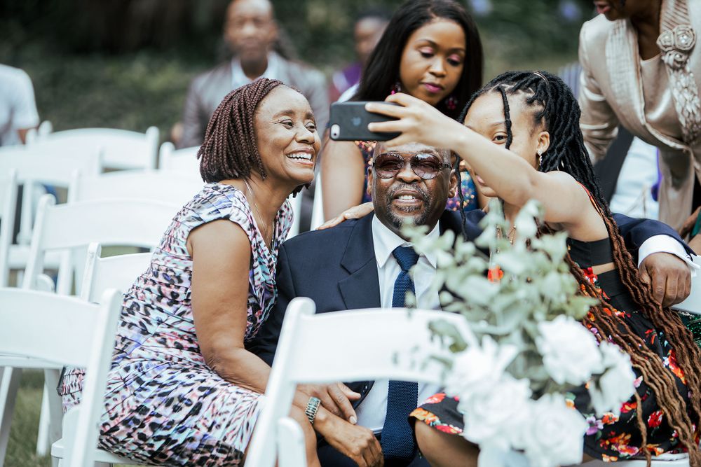 Wedding guests laughing and socializing in white chairs during outdoor ceremony.
