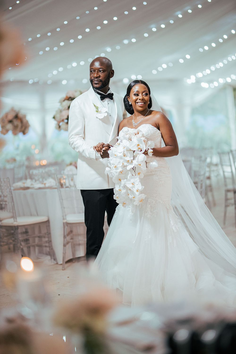 A wedding couple stands together in a luxuriously decorated reception space with string lights overhead.