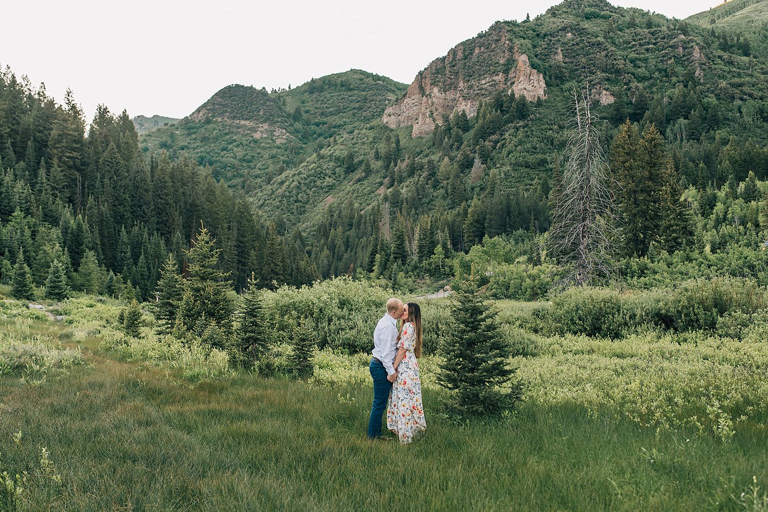 Couple standing in a mountain meadow surrounded by evergreen forest and rocky peaks.