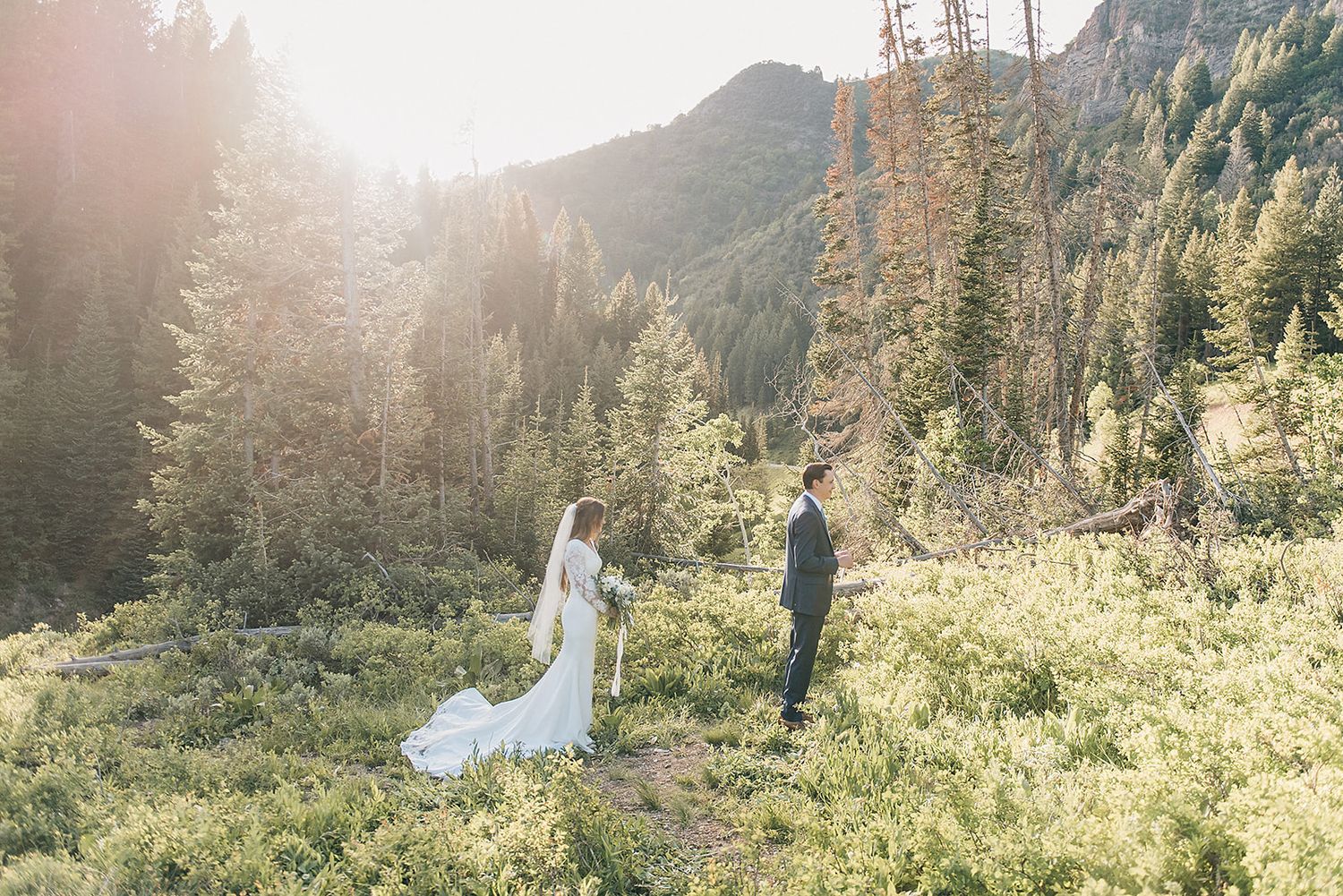 Wedding couple stands in a sunlit mountain meadow surrounded by pine forest.