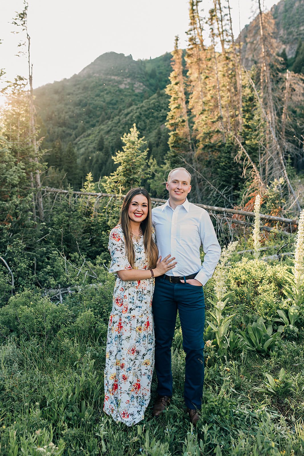 Engaged couple posing in floral dress and casual attire against mountain wilderness backdrop at sunset.