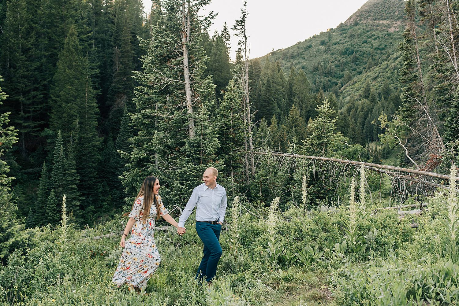 Couple walking hand in hand through lush mountain wilderness during engagement photo session.
