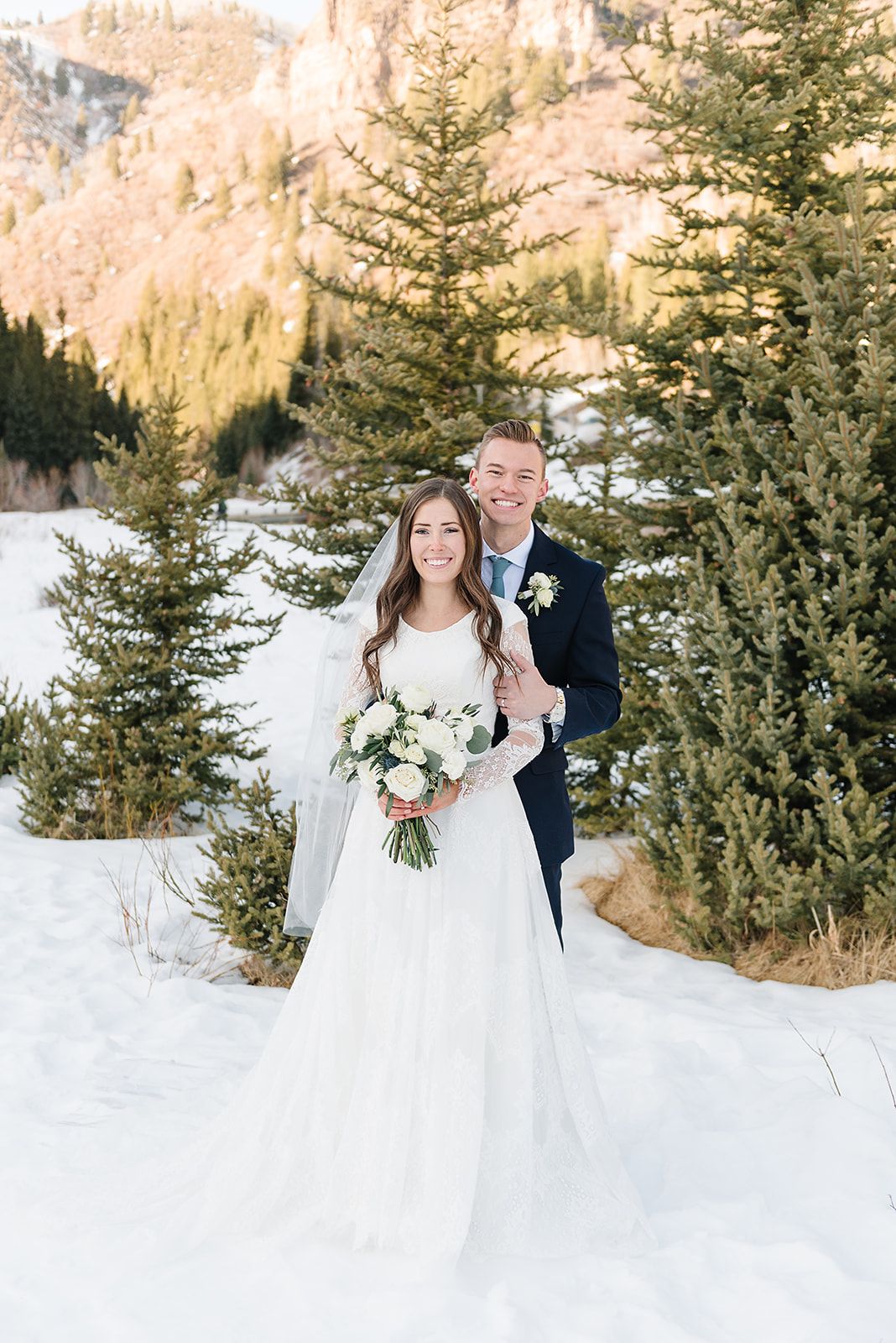 Elegant winter wedding portrait with snowy mountains and evergreen trees creating a stunning backdrop.