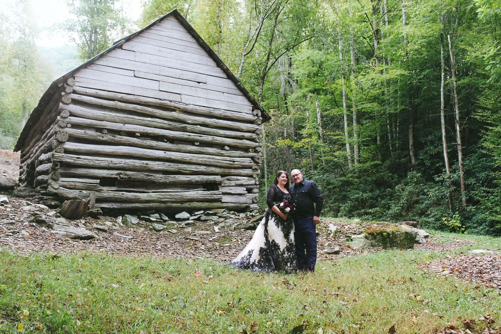 A couple poses in front of rustic log cabin in woodland setting.