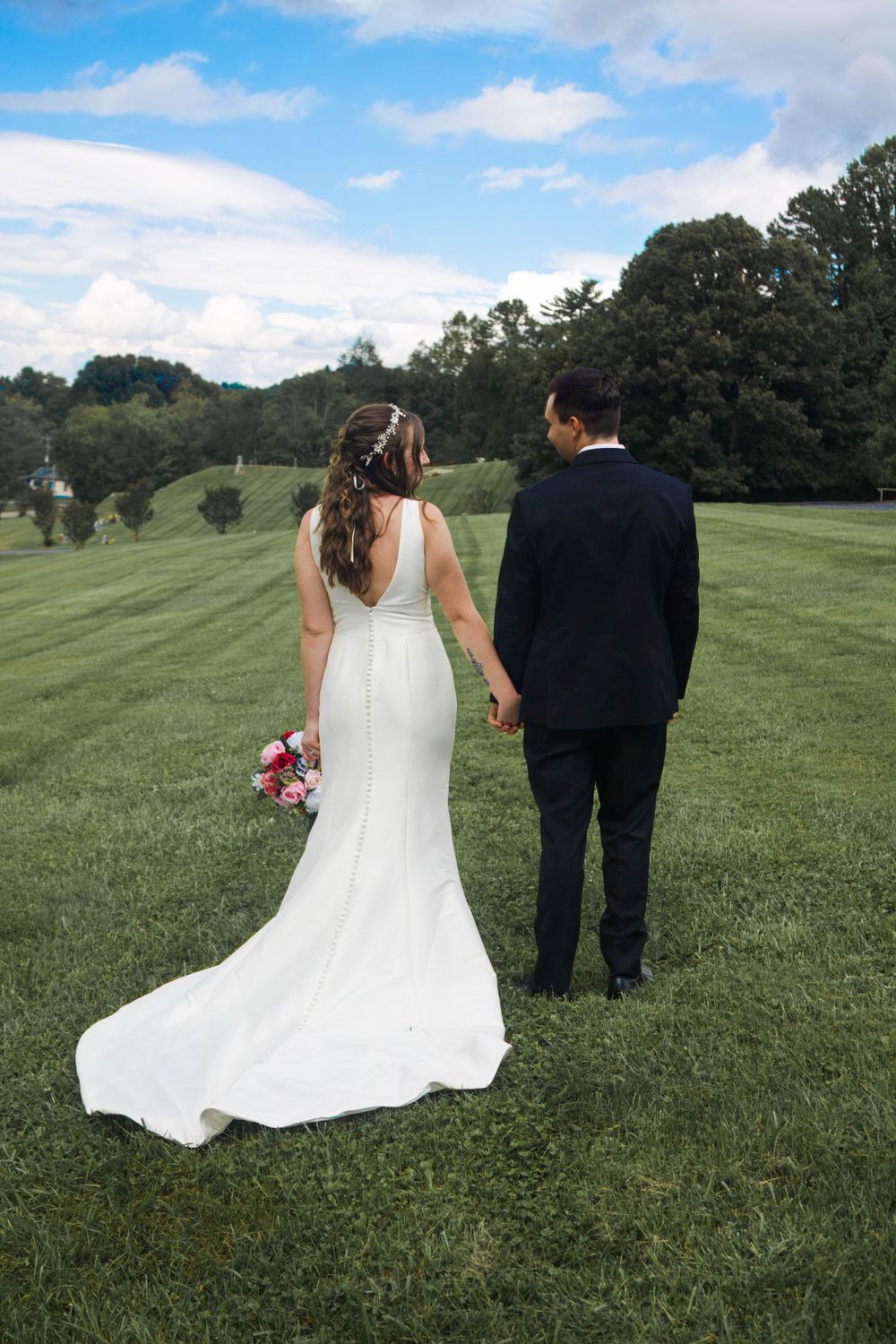 Newlyweds walk hand in hand across a scenic hilltop with bouquet and flowing dress.