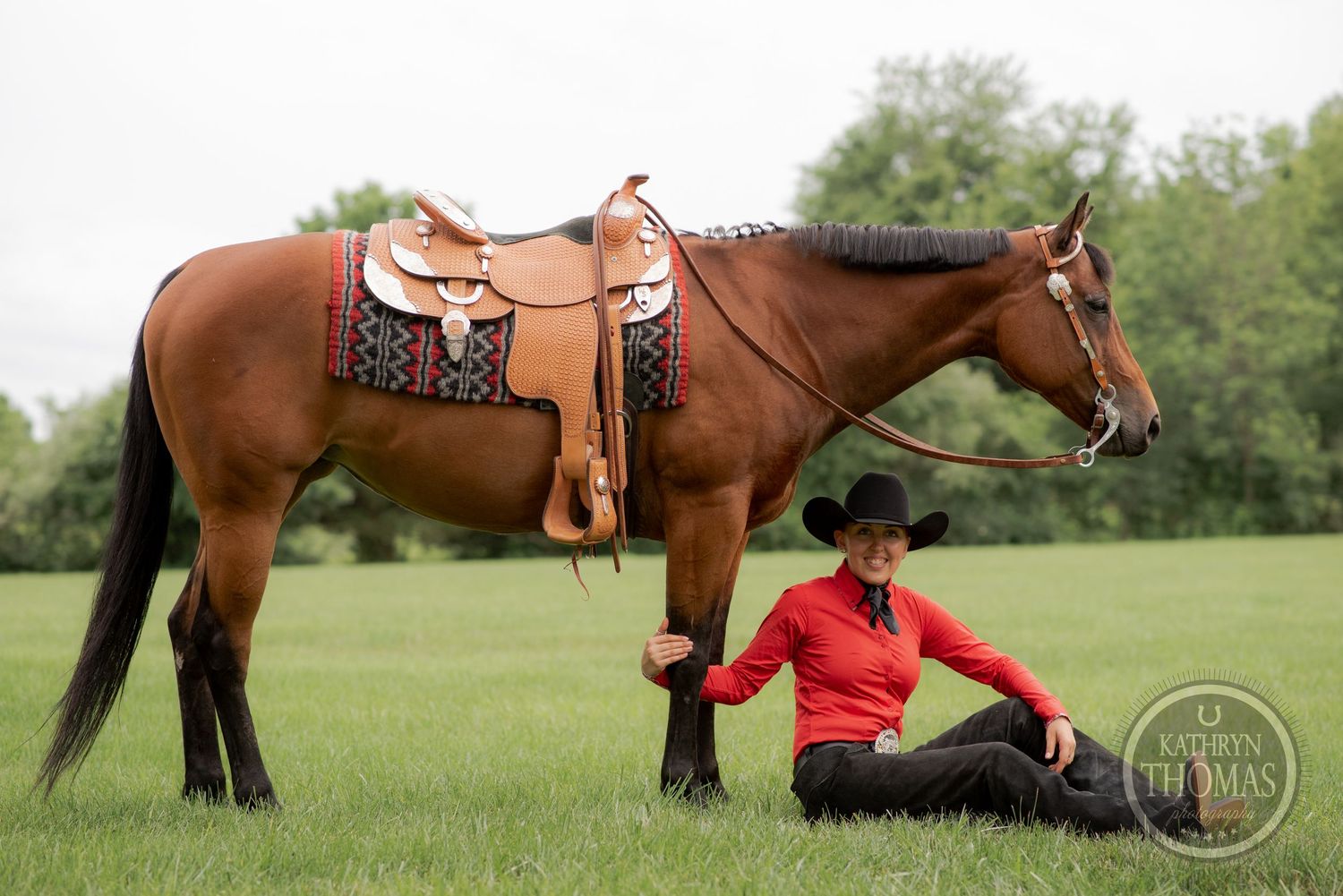 Brown horse with decorative western saddle while handler rests in grass.