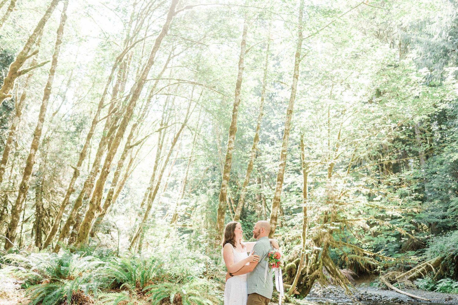 A couple in forest setting embrace among tall trees and lush vegetation.